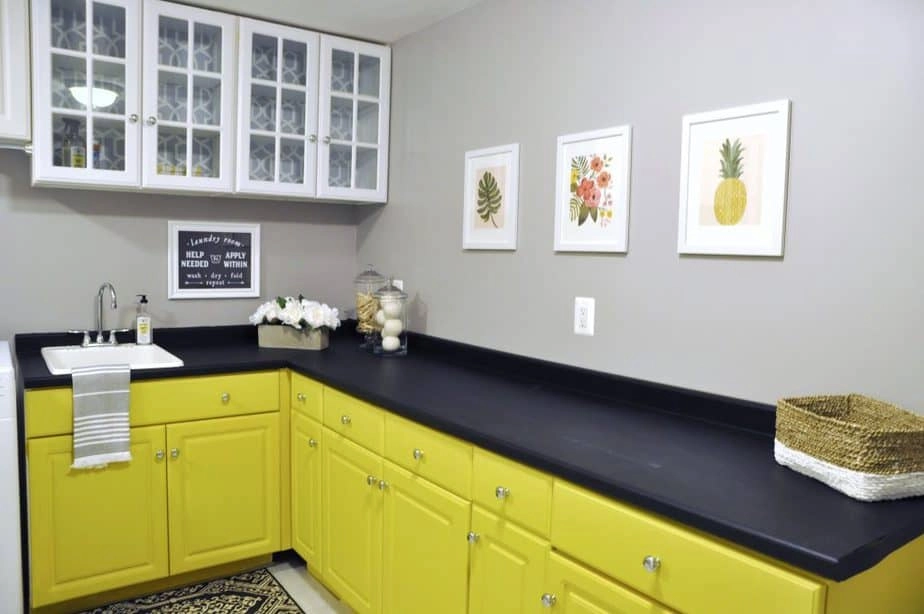 Updated laundry room cabinets painted bright yellow, black countertops painted with chalk paint, and tropical wall art hung on the walls.