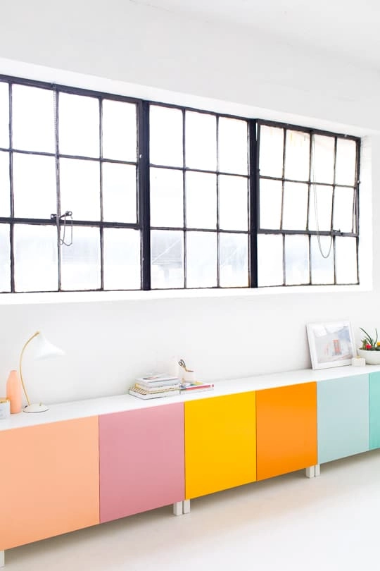 A set of 3 IKEA Besta modular cabinets sits along a white wall. Each cabinet door is painted with a bright color - pink, purple, yellow, orange, and blue.