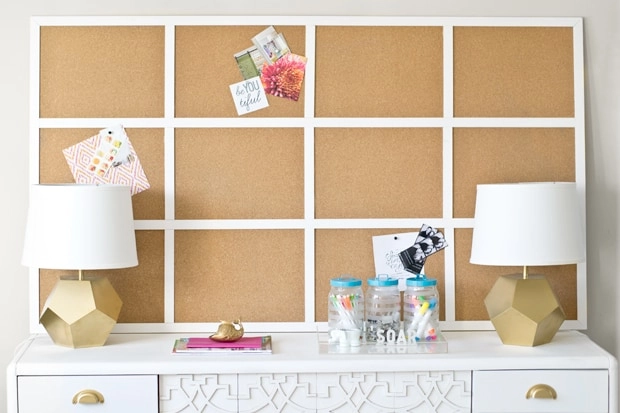 A 12-grid cork board command center with white wood frame sits on top of a white dresser with gold and white geometric lamps,.