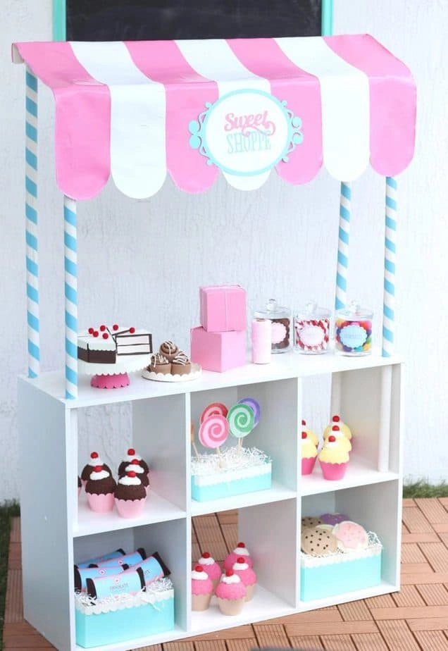 This DIY kids sweet shoppe play station is made with an IKEA Kallaz shelf turned on it's side. Play cakes and desserts fill the shelves and a DIY pink and white shop canopy is made with paper and cardboard tubes.