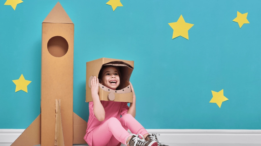 A little girl plays astronaut with a cardboard rocket and space helmet, sitting on the floor in front of a blue wall decorated with yellow paper stars.