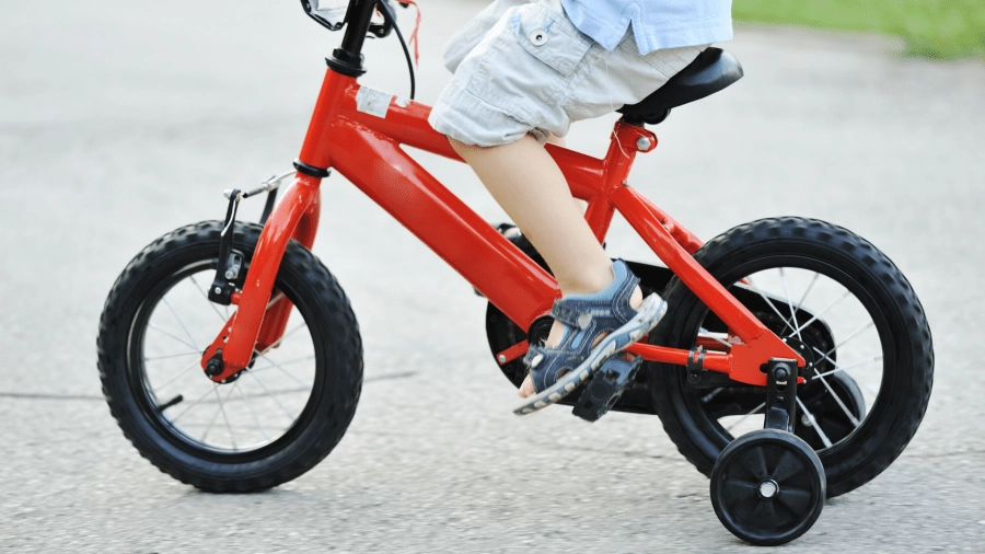 A little boy rides on a red bicycle with training wheels around his neighborhood.