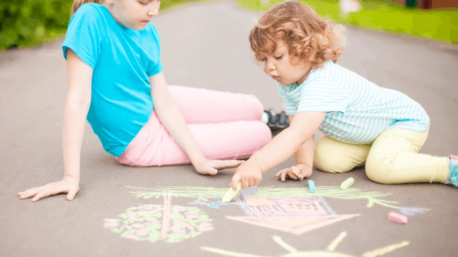 Two young girls color on a sidewalk with pastel colored chalk, drawing a house and tree with flowers.