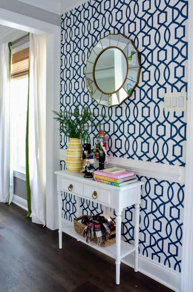 One wall in our entryway foyer with blue and white patterned wallpaper and a round sunburst mirror. A small white end table sits against the wall, decorated with books, a vase, and storage basket.
