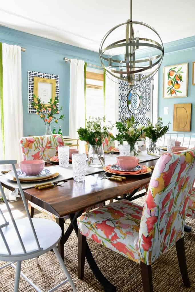 Another look at our dining room makeover: the dining room table is set with gold, white, and coral dinner ware, crystal glasses, and colorful abstract fabric chair covers.