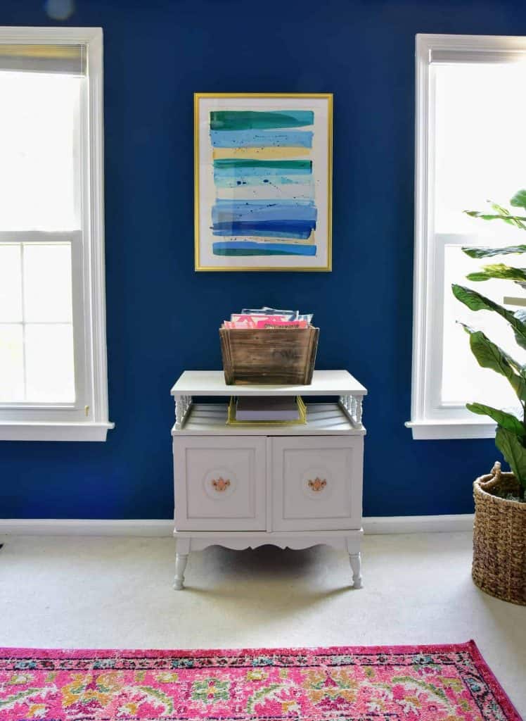 A thrifted storage cabinet in our home office - we painted it white for an upgrade. It sits against a navy blue wall, under an abstract blue striped art print.