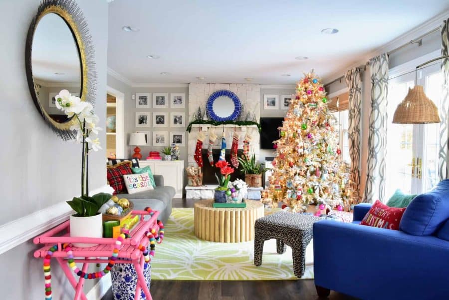 A colorful living room decorated for Christmas - a large faux white Christmas tree, stockings hanging on the mantle, multi-colored garland stands on a small pink side table, and festive Christmas throw pillows on the couch and chairs.