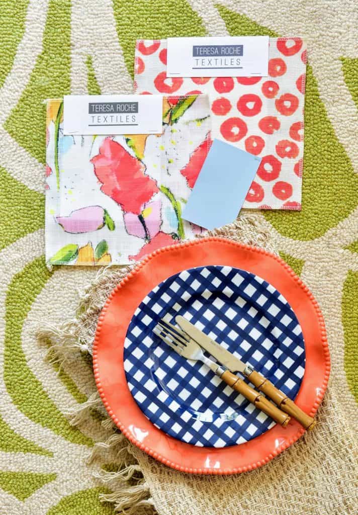 Colorful Spring fabric swatches sit on a green and white patterned rug next to a table setting of a coral charger plate, blue and white hatch patterned dinner plate, and bamboo-handled cutlery.